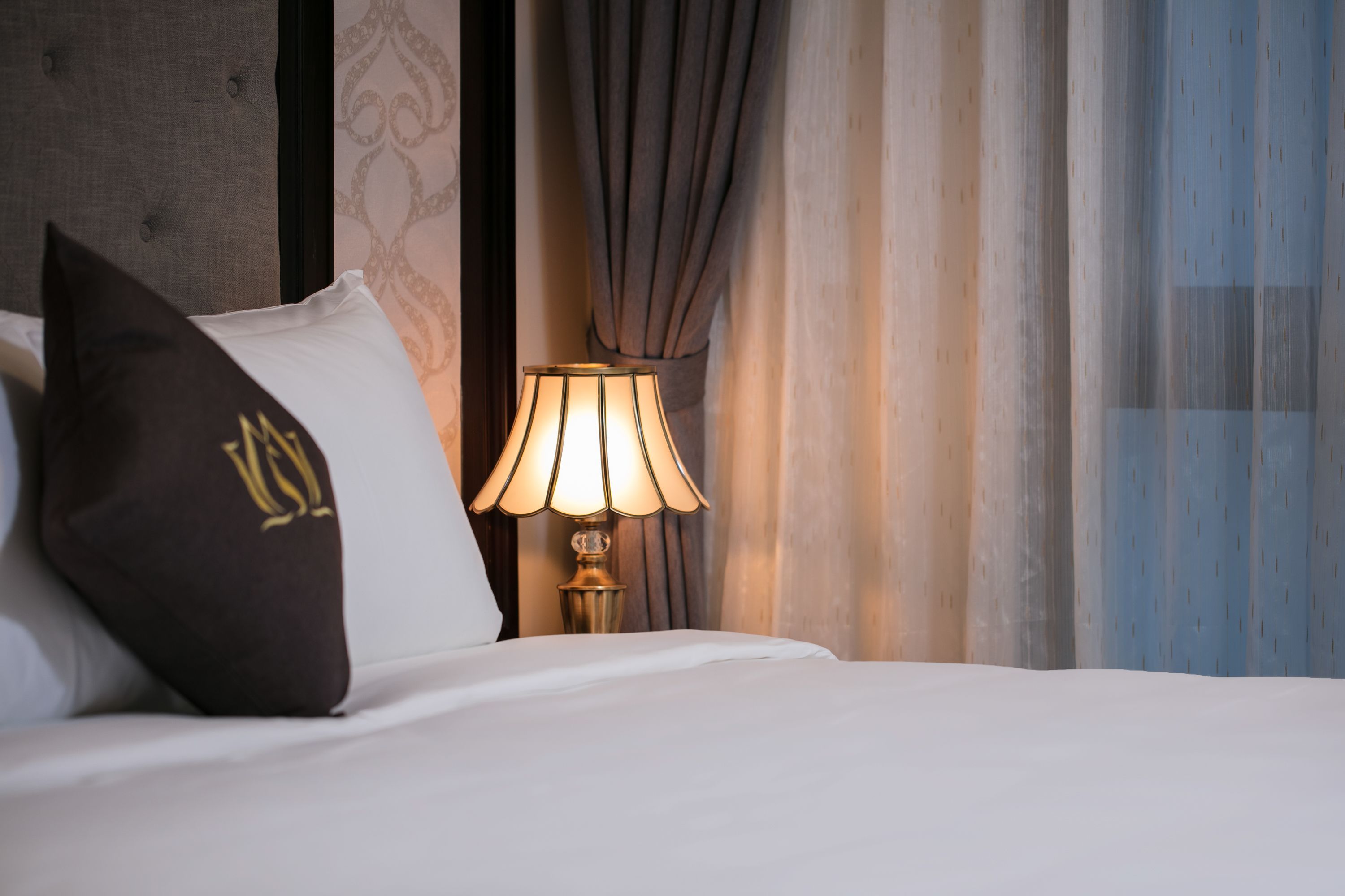 A neatly made bed with white linens and a dark brown pillow with a gold emblem. A table lamp with a beige lampshade is placed beside the bed.