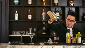 A bar setting with a bartender pouring a drink into a martini glass, a bottle of liquor, and a bottle of juice on the counter. - Sen Grand Hotel & Spa managed by Sen Group (Hanoi)