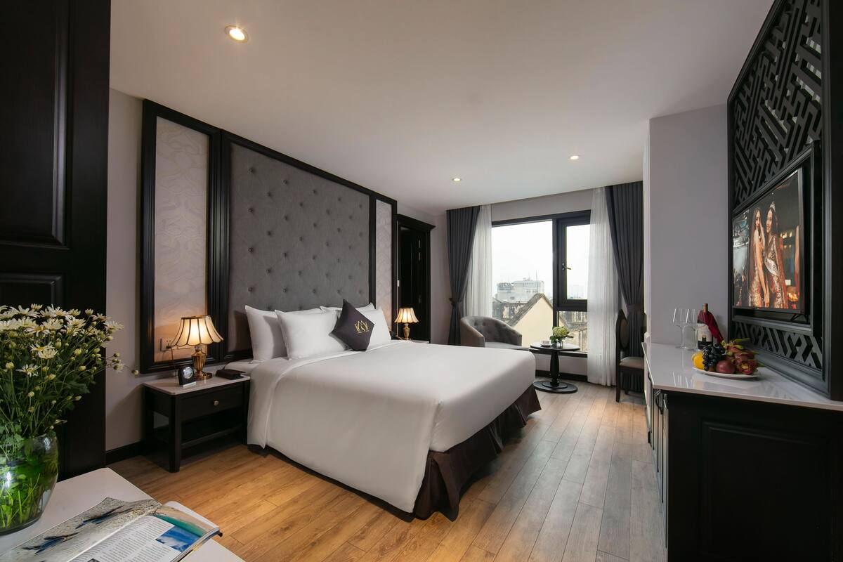 Een hotelkamer met een groot bed, een nachtkastje met een lamp, een stoel, een bureau met een bloevas en uitzicht op de stad door het raam.