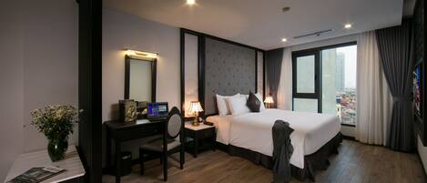 A hotel room with a large bed, a desk with a computer, a chair, a small table with a vase of flowers, and a window with curtains.