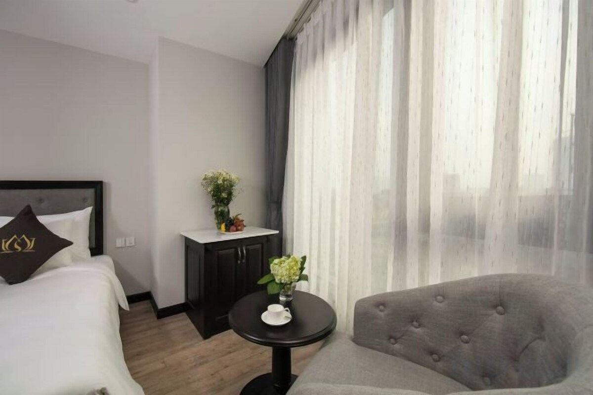 A hotel room with a bed, a small table with a vase of flowers, a grey armchair, and a large window with sheer curtains.