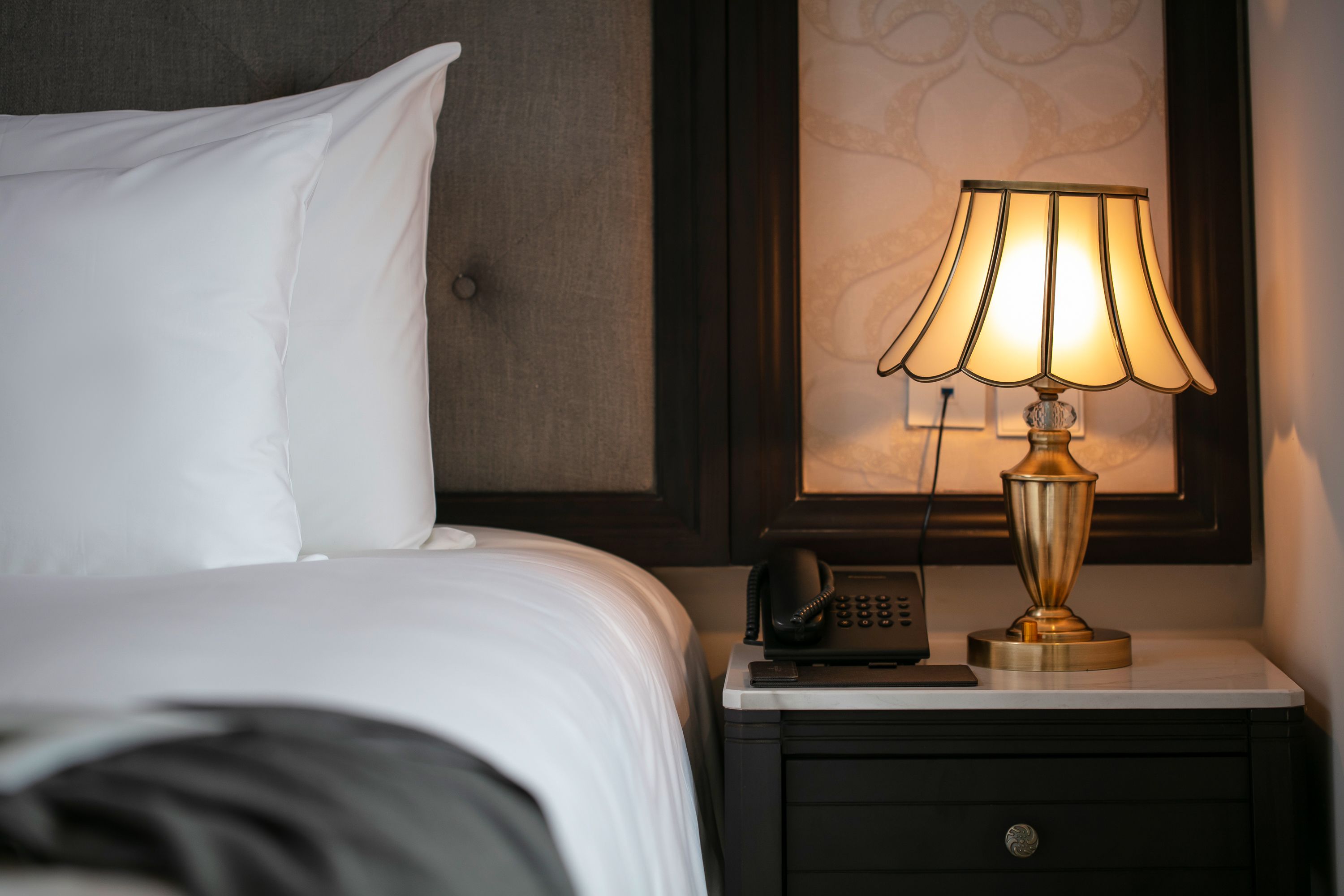 A neatly made bed with white linens, a bedside table with a lamp, a telephone, and a small drawer.