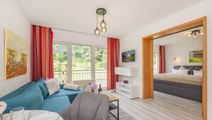 A modern living room with a sofa, a dining table with plates and glasses, a TV, and a bed with a view. - Cozy vacation apartment in Heringsdorf just 200 m from the Baltic Sea beach (Seebad Heringsdorf)