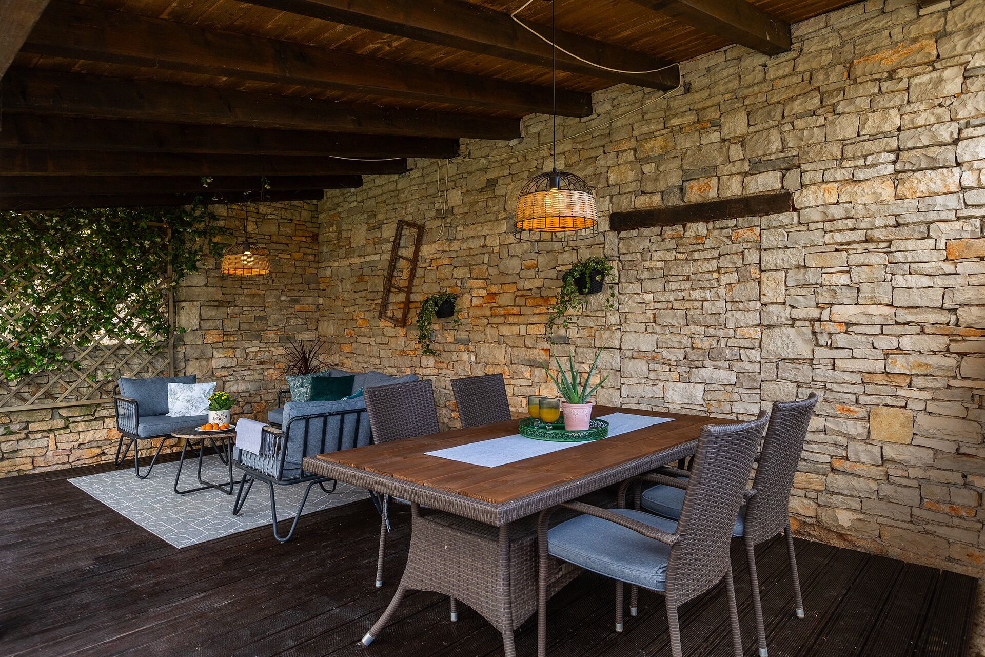 Interior rústico con pared de piedra, una mesa de madera, sillas de mimbre, un candelabro colgante y una escalera.
