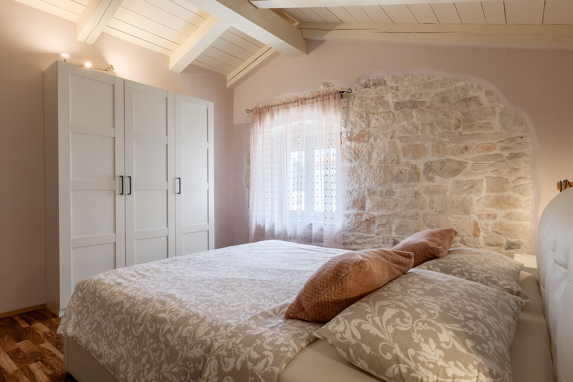 A bedroom with a stone wall, a bed with patterned bedding, and a wardrobe.