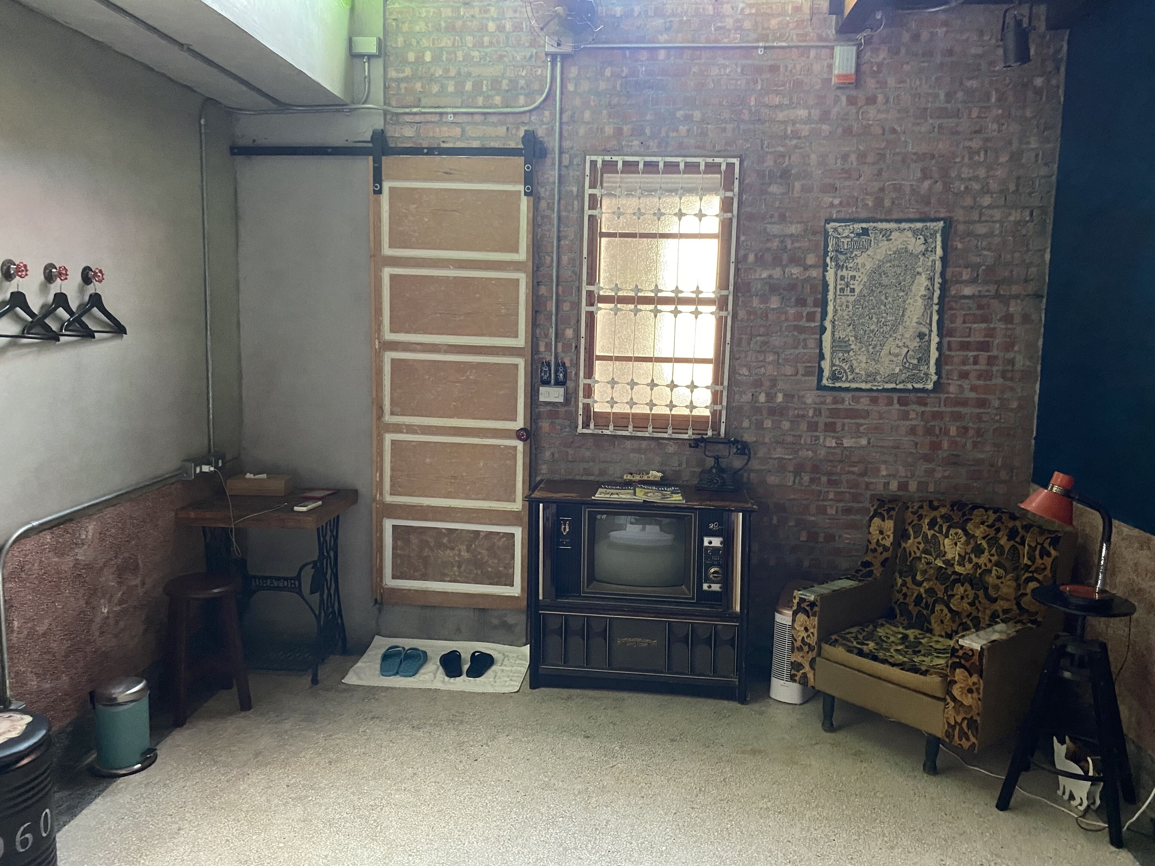 A compact living space with a brick wall, a vintage television, a patterned armchair, and a small table with a lamp.