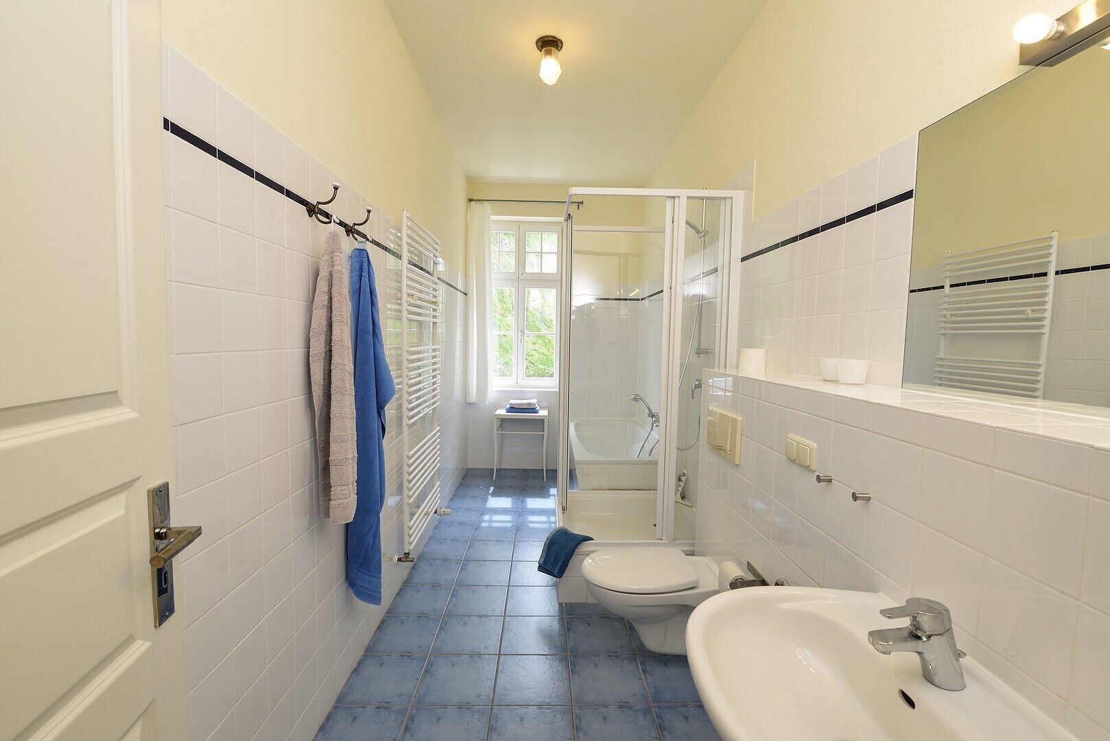 A narrow bathroom with white tiles, a toilet, a sink, a shower, and a towel rack.