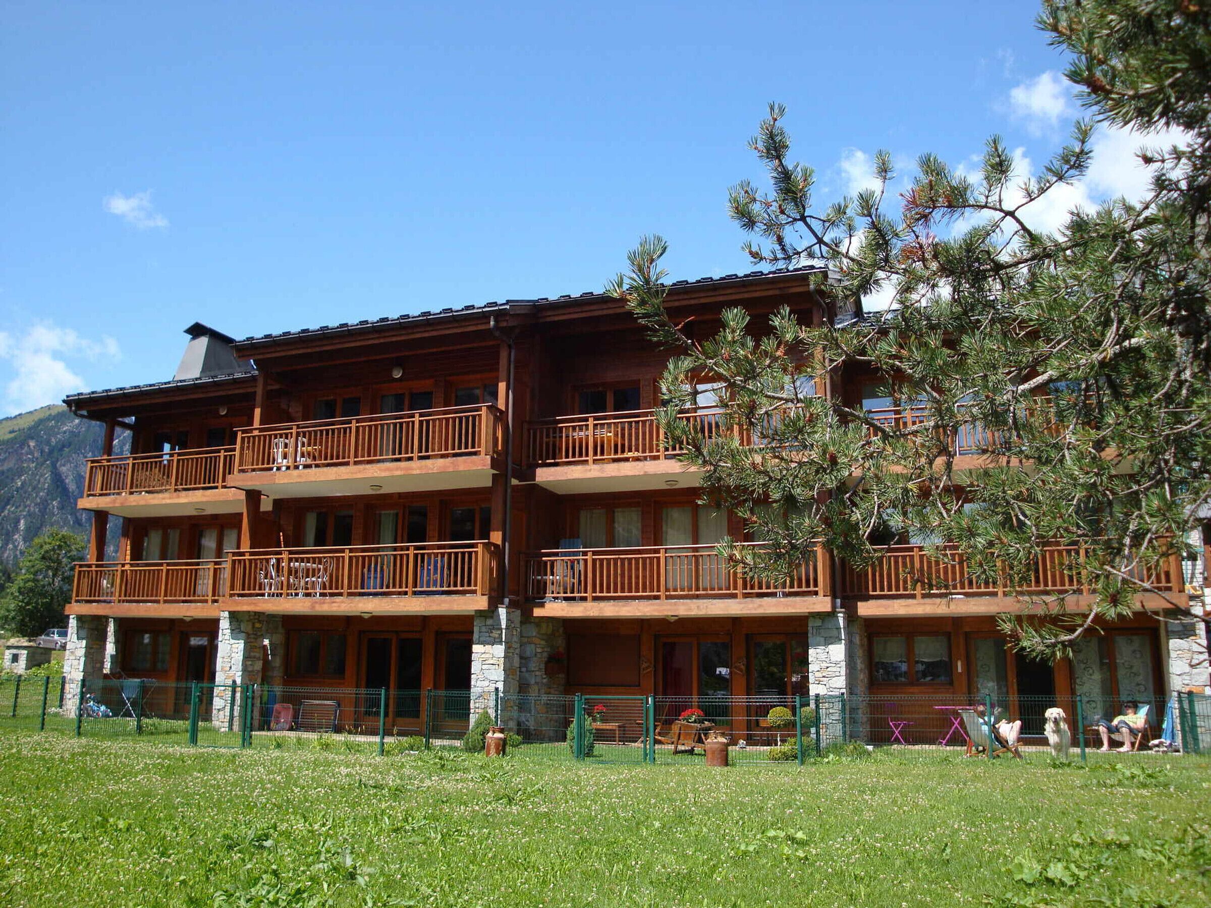 A multi-story wooden building with balconies, stone foundation, and a grassy front yard.