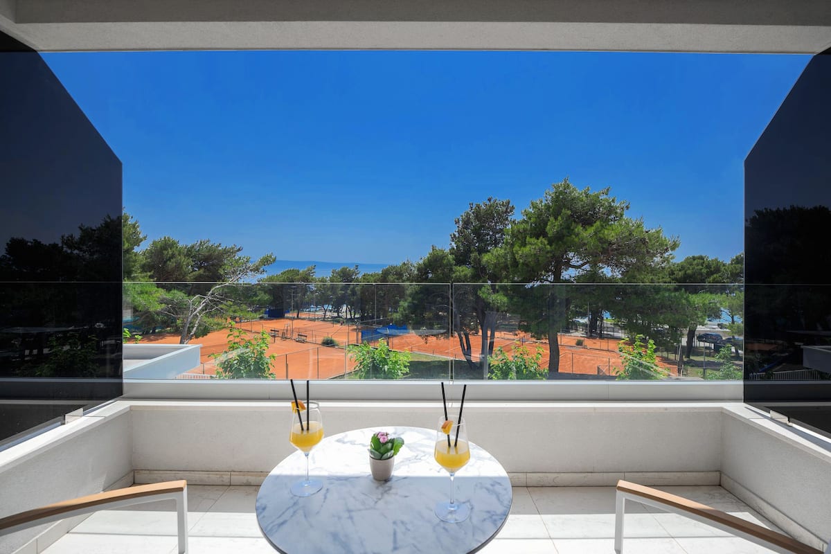 A balcony with a clear view of a tennis court, a table with two drinks, and a small plant.