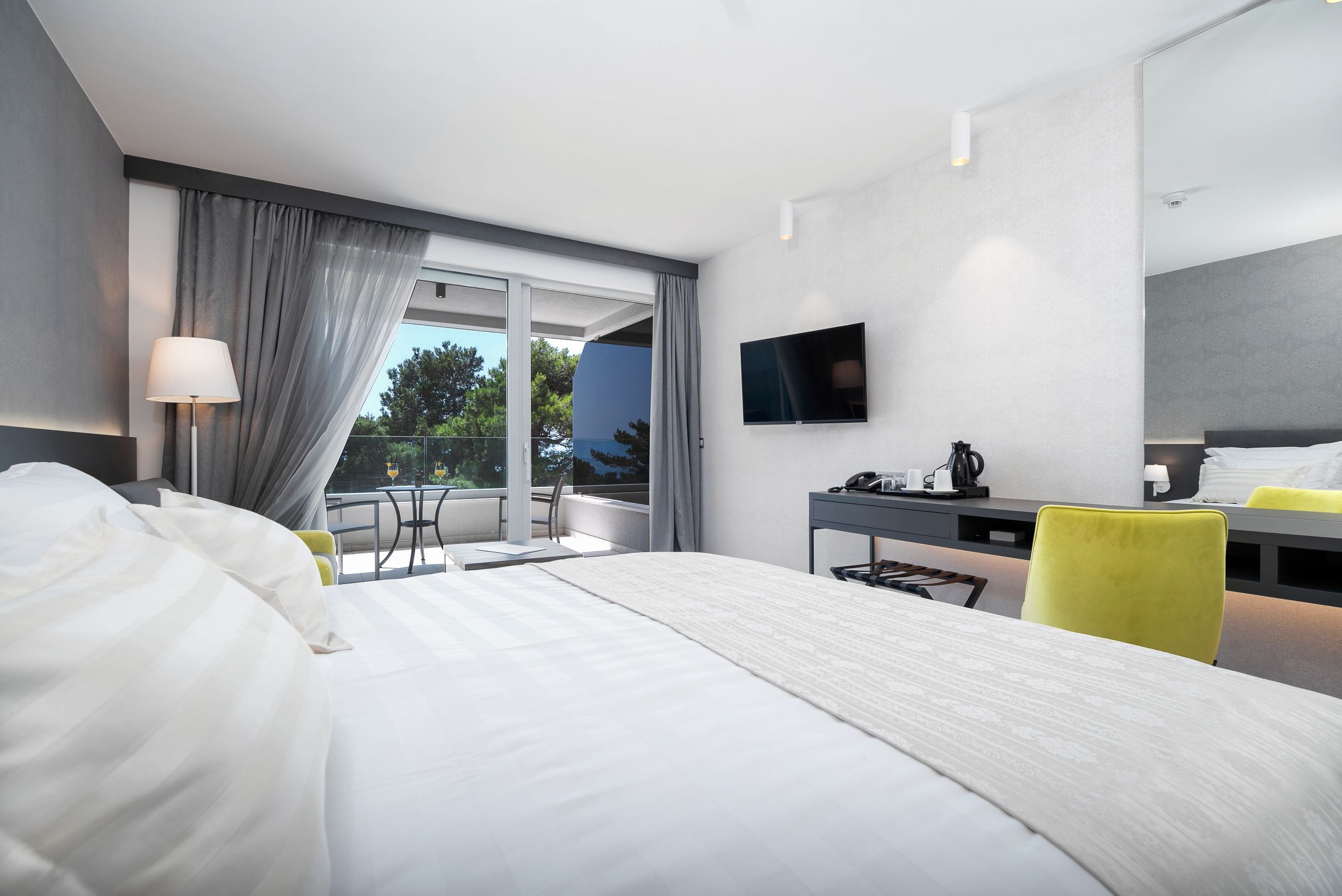 Ein modernes Hotelzimmer mit einem großen Bett, einem Schreibtisch mit Stuhl, einem an der Wand befestigten Fernseher und einem Balkon mit Blick auf Bäume.