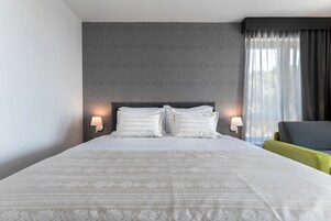 Een hotelkamer met een groot bed, twee bedlampen, een grijs hoofdeinde, een grijze fauteuil en een raam met gordijnen.