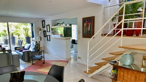 Sala de estar moderna com mesa de jantar de vidro, cadeiras pretas, escada e vista para o exterior.