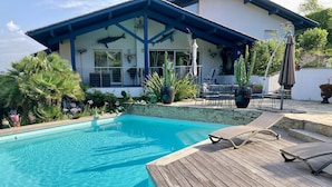 Piscina com deck de madeira, cercada por plantas tropicais e uma casa com telhado azul e decoração de tubarão.