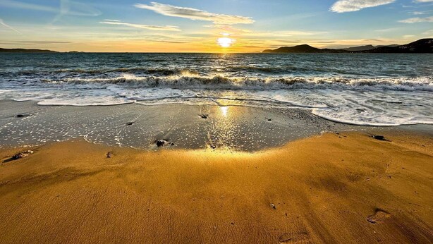 Una playa con un atardecer sobre el océano, una orilla arenosa y montañas distantes.