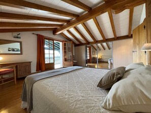 A bedroom with a large bed, wooden beams, a desk, and a sliding glass door leading to a balcony.