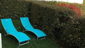Duas cadeiras de descanso azuis numa área gramada, com um muro de sebe e um edifício ao fundo.