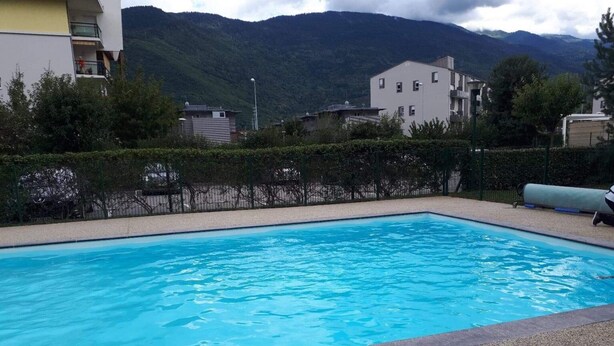 A rectangular swimming pool with clear blue water, surrounded by a pebble border. In the background, there are buildings with balconies and a mountainous landscape. - Lovely T2 garden apartment with sheltered terrace swimming pool (summer) Fibre  (Albertville)