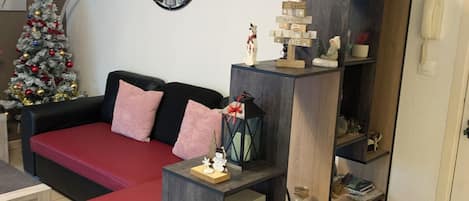 A living room with a red sofa, a wooden coffee table, a clock, a Christmas tree, and a bookshelf with various items.