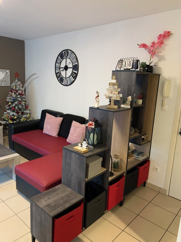 A living room with a red sofa, a wooden coffee table, a clock, a Christmas tree, and a bookshelf with various items.