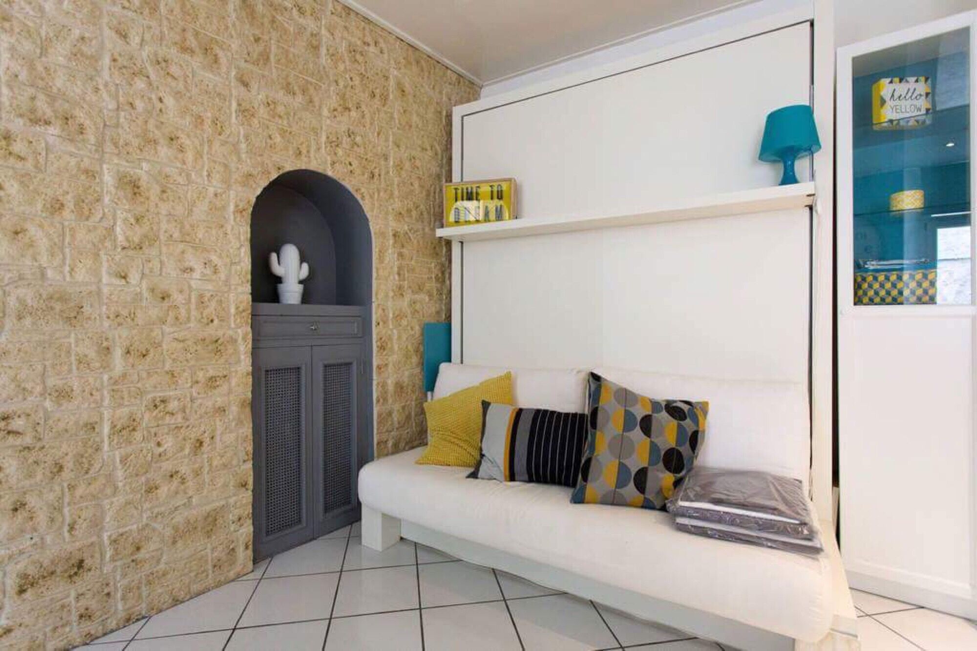 Una habitación con pared de piedra, un sofá blanco con cojines estampados, una mesita con una lámpara y una estantería con objetos decorativos.