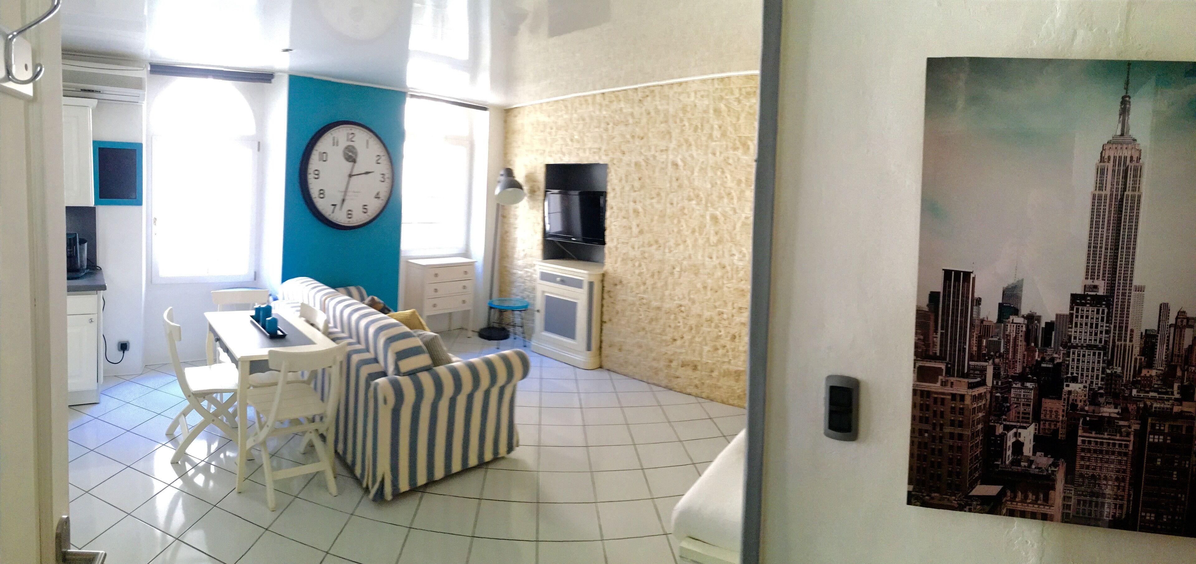 Un salon avec un canapé, une table à manger et des chaises. Il y a une horloge au mur et un tableau représentant un paysage urbain.