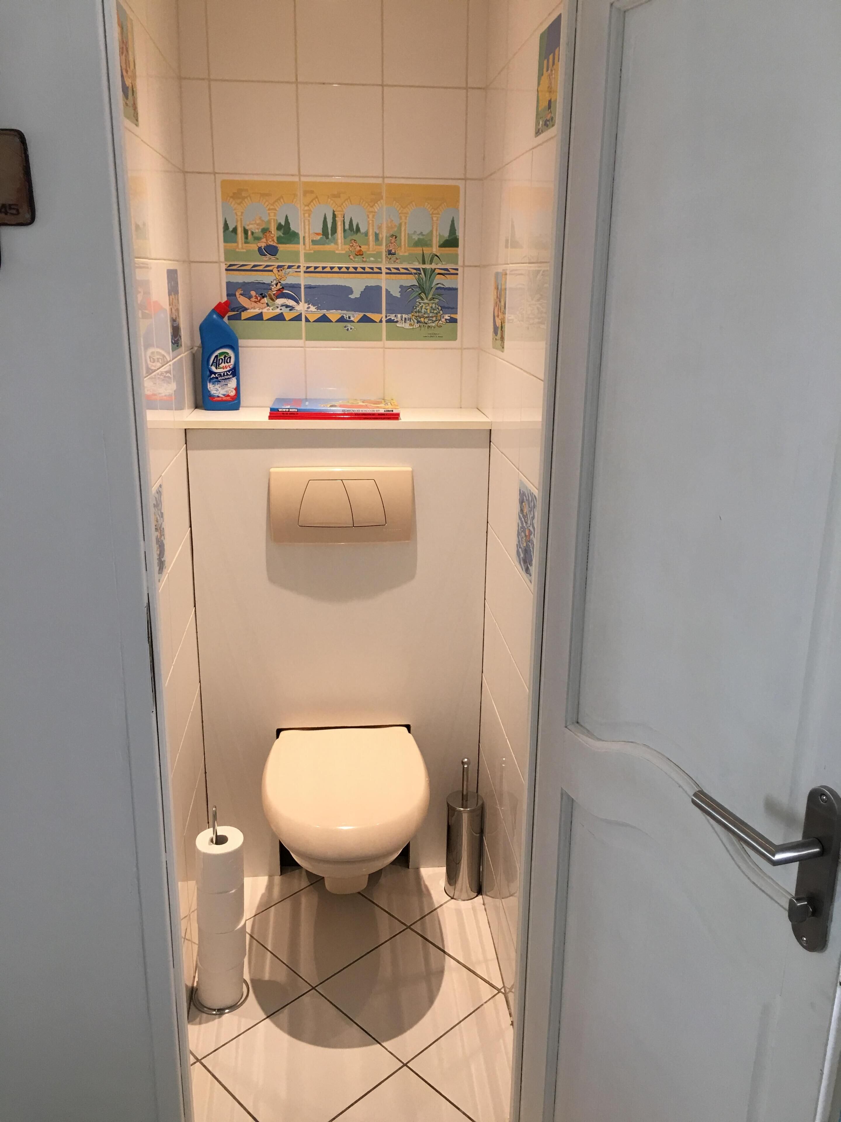 A small bathroom with a toilet, a toilet paper holder, and a shelf with a bottle of cleaning product.