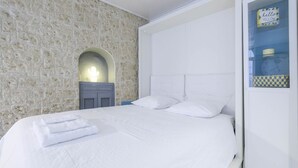 Una cama bien hecha con sábanas y almohadas blancas, una pared de piedra con un detalle decorativo de cactus y un cabecero blanco.