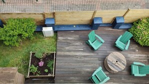 A wooden deck with green chairs and a round table, surrounded by raised garden beds and a brick pathway. - Villa Azious, 10 persons, beach side in Wimereux with parking possibility (Wimereux)