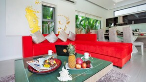 A modern living room with a red sofa, a glass coffee table with fruits and magazines, and a kitchen in the background. - Isawan 2BR Salt Pool Villa in Baannaraya (Rawai)