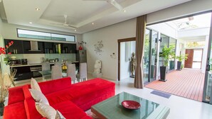 A modern living room with a red sofa, a glass coffee table, and a view of the outdoor patio area. - Isawan 2BR Salt Pool Villa in Baannaraya (Rawai)