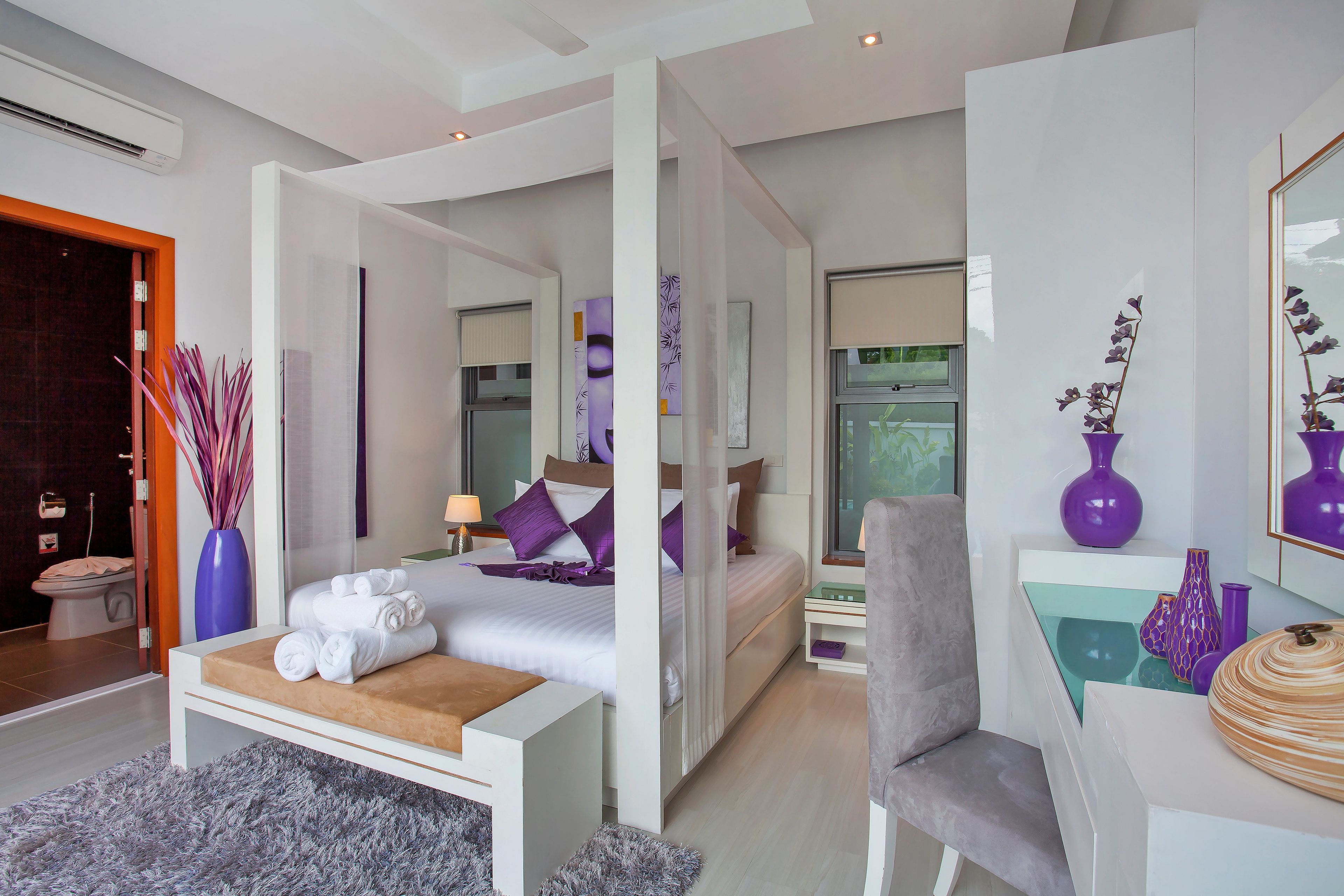 A modern hotel room with a large bed, a desk, and a chair. There is a bathroom visible through an open door.