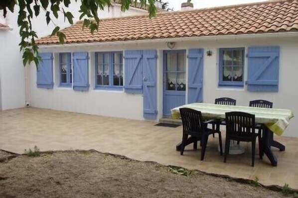 Un bâtiment blanc avec des volets bleus, un toit en tuiles et une table avec des chaises à l’extérieur.