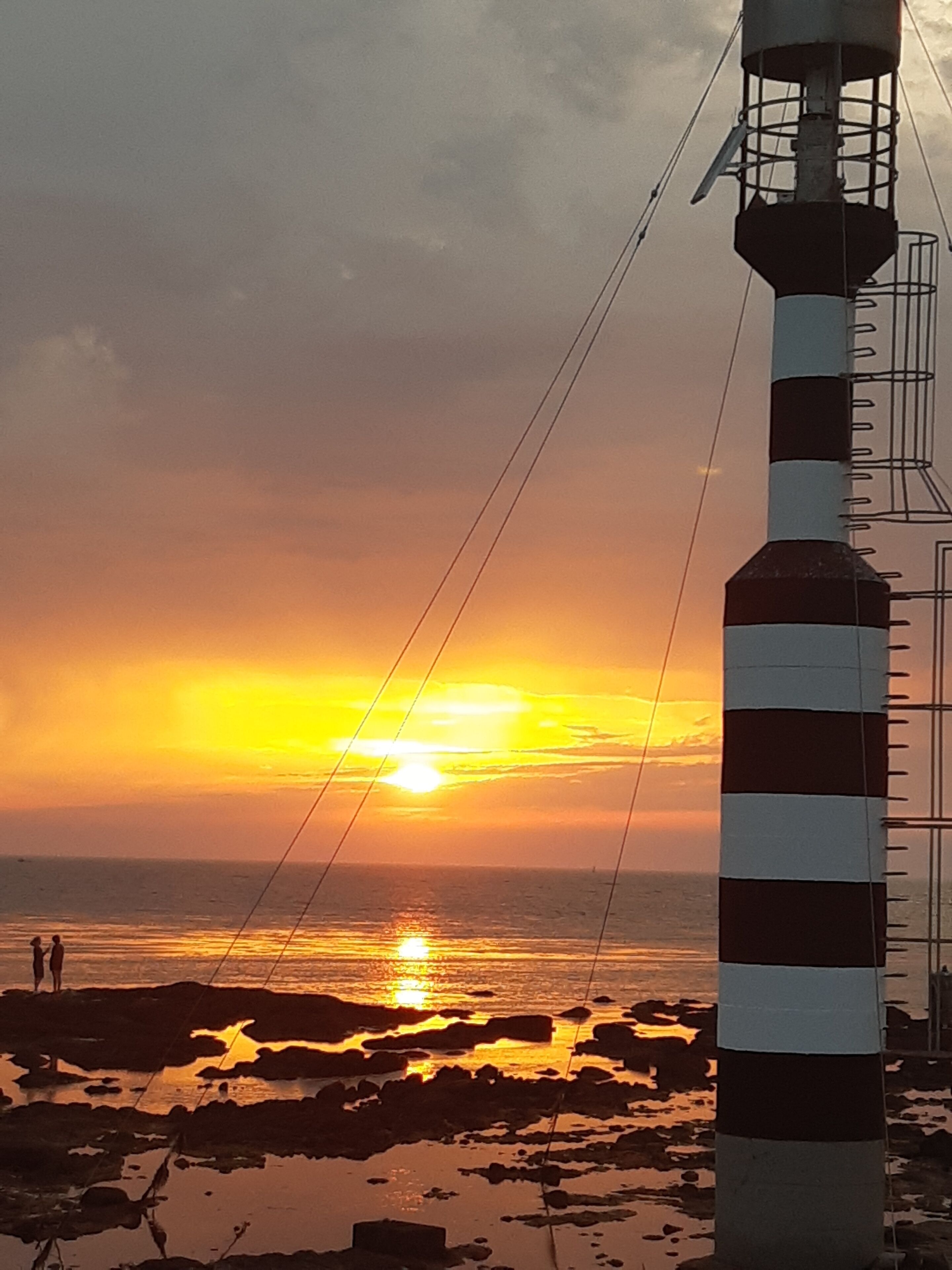 Een vuurtoren met een gestreept patroon, uitkijkend over een rotsachtige kustlijn met een ondergaande zon.