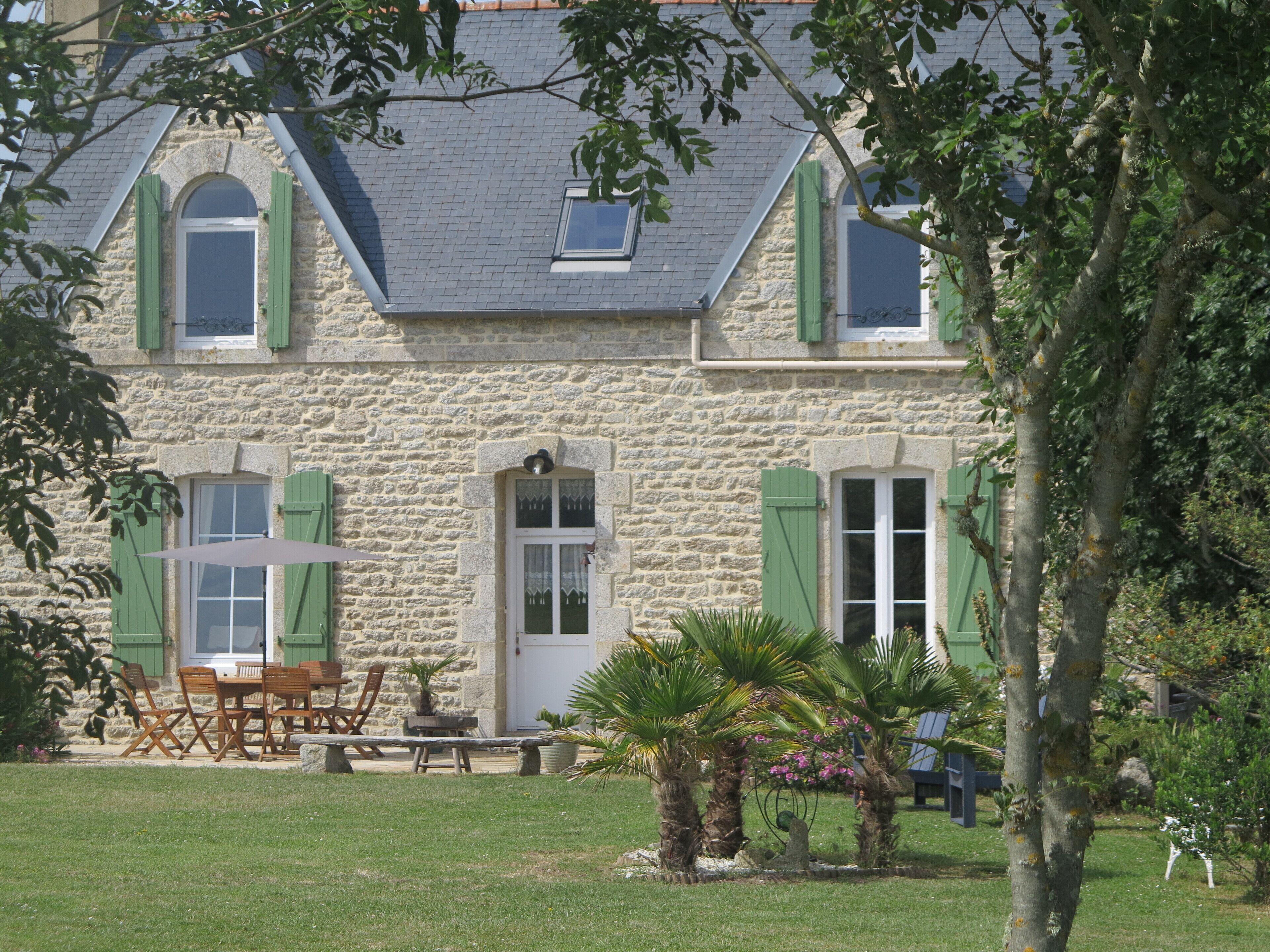 A stone building with green shutters, a patio with outdoor furniture, and a small garden with palm trees.