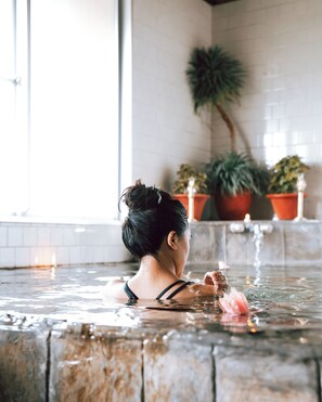Une personne dans un bain à remous, avec un mur carrelé et des plantes en pot en arrière-plan.