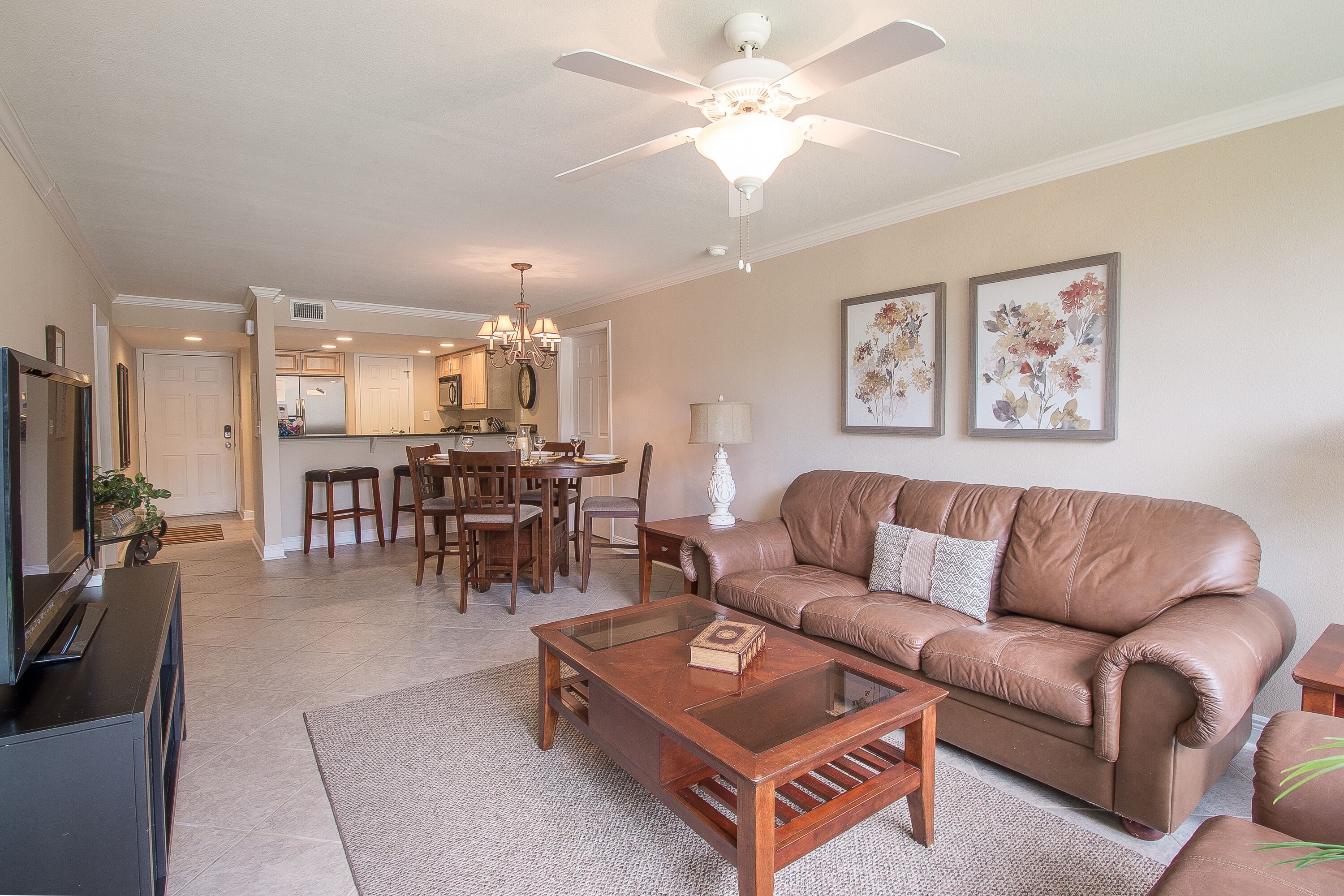 A living room with a brown leather sofa, a glass coffee table, and a TV stand. There is a dining area with a chandelier and a kitchen in the background.