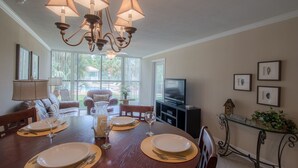 A dining area with a wooden table set for four, a chandelier, and a television in the background. - Let The Waves Hit Your Feet & Sand Be Your Seat! Step Right Out To The Pool! (Biloxi)