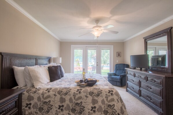 A bedroom with a large bed, a ceiling fan, a dresser, a television, and a view of the outdoors through the door.