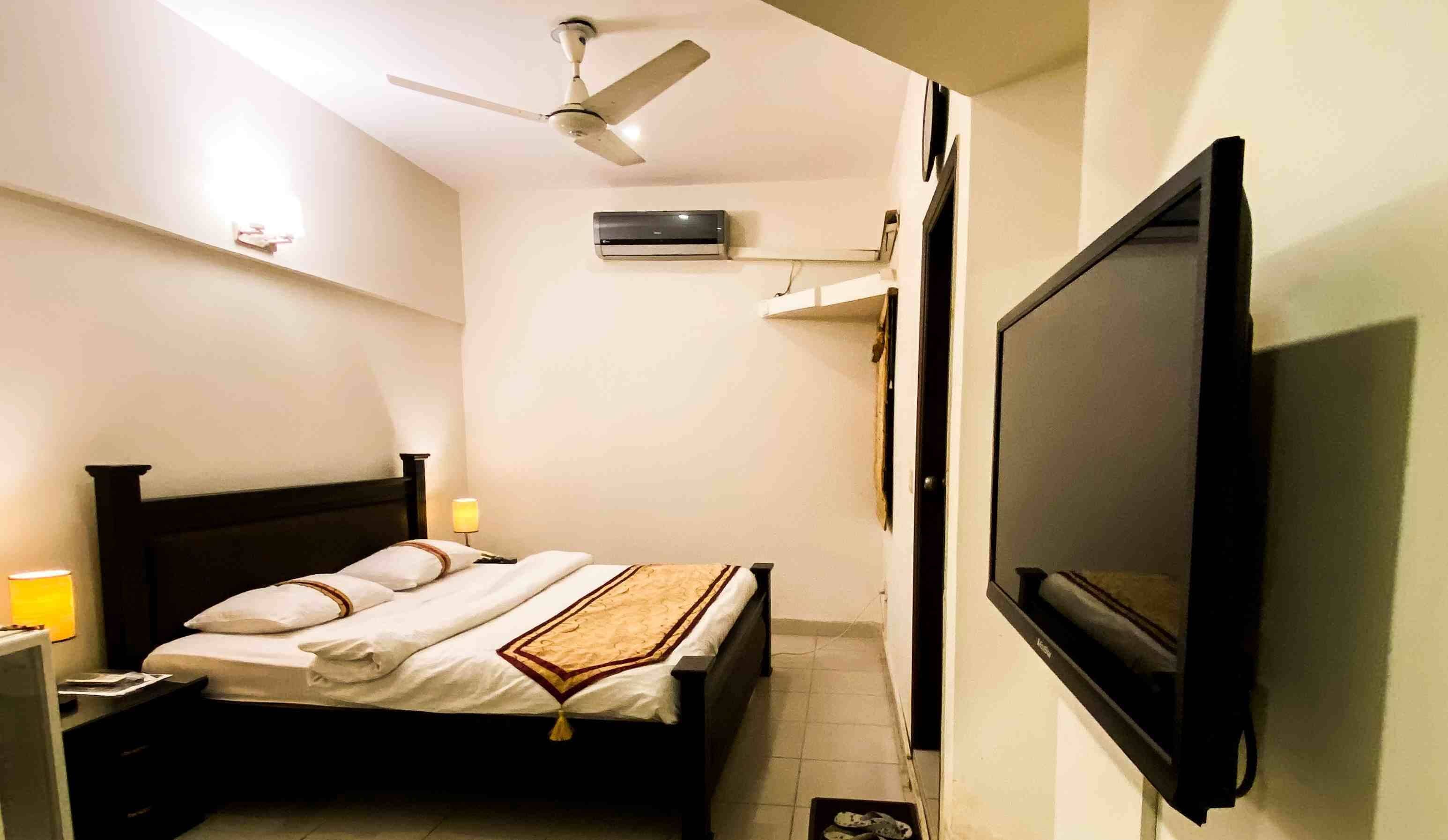 Un dormitorio con cama, lámparas de noche, ventilador de techo, unidad de aire acondicionado y televisor montado.