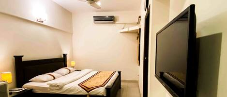 Un dormitorio con cama, lámparas de noche, ventilador de techo, unidad de aire acondicionado y televisor montado.