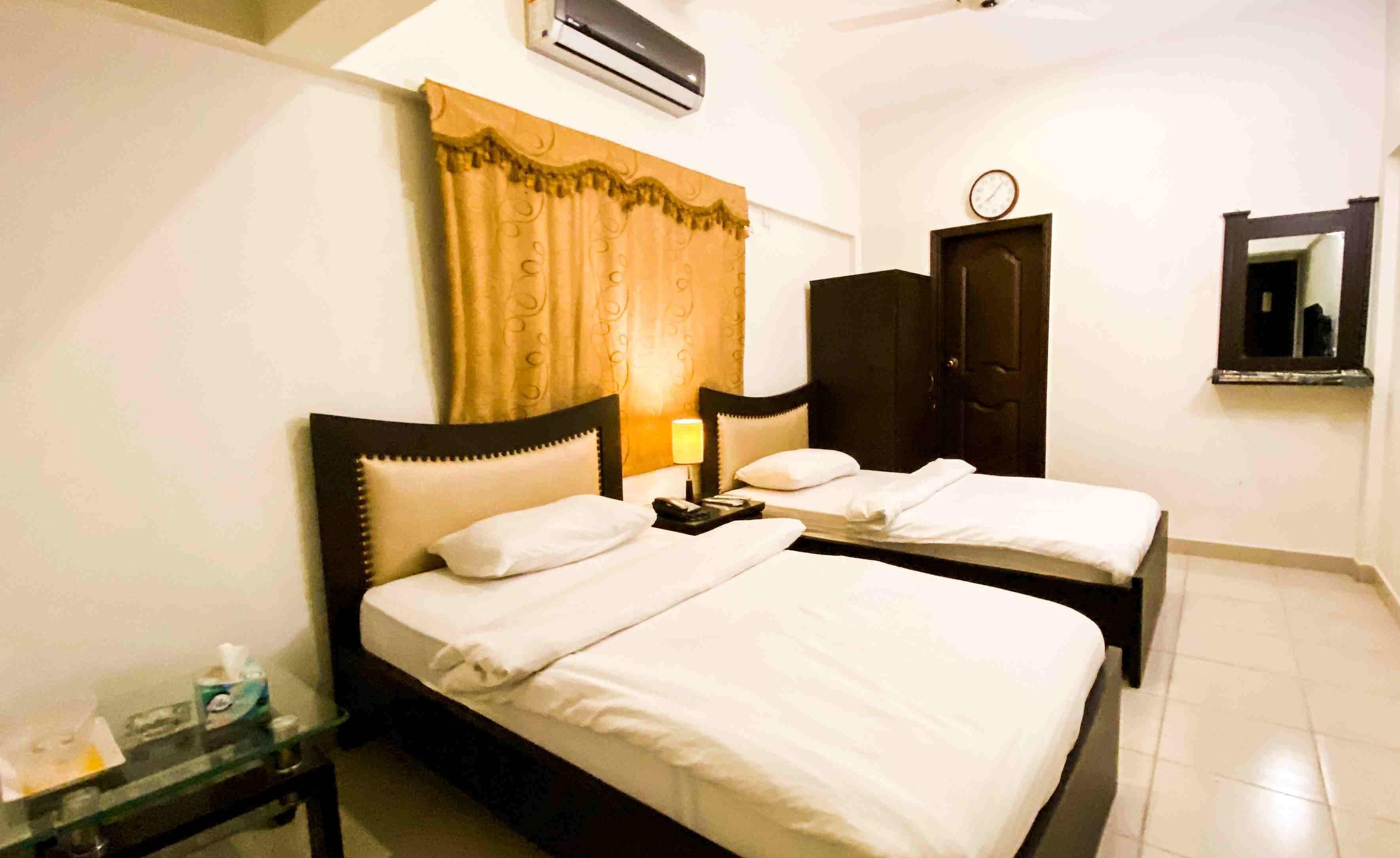 Quarto de hotel com duas camas, uma mesa de cabeceira com abajur, um relógio na parede e um espelho.