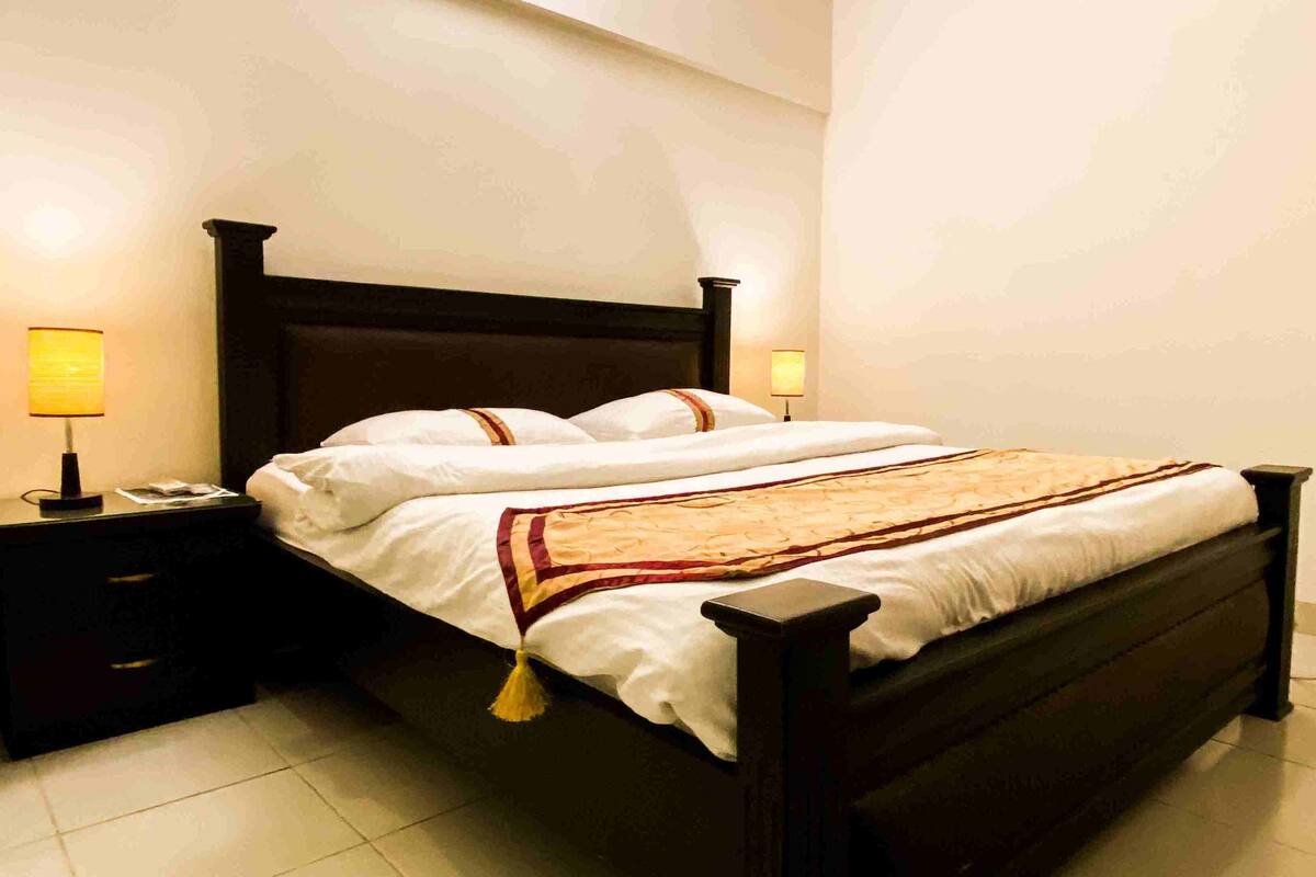 A neatly made bed with white linens and a decorative throw, flanked by two bedside tables with lamps, in a room with plain walls and tiled flooring.