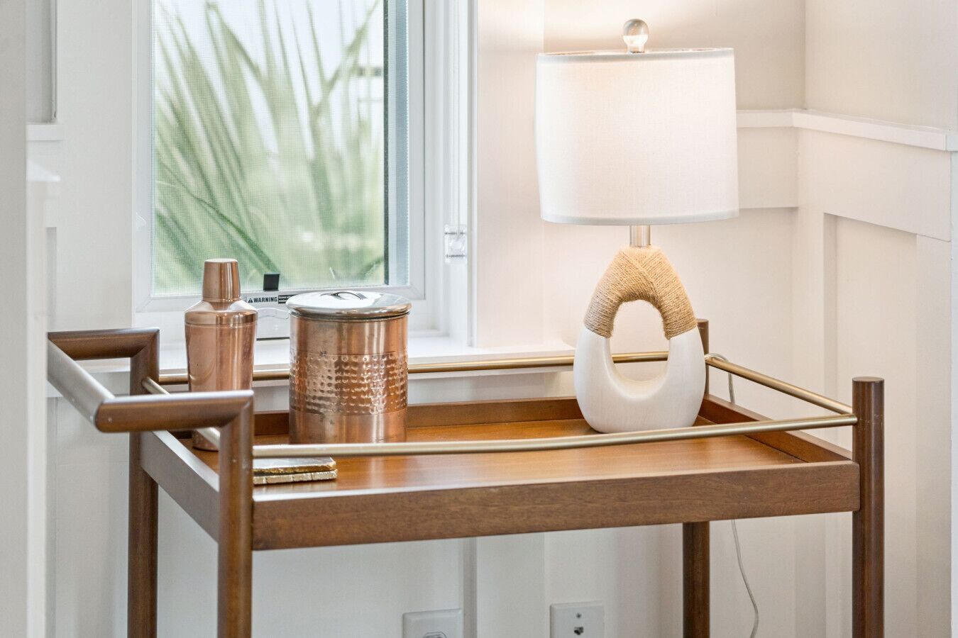 A wooden side table with a lamp and decorative items by a window.