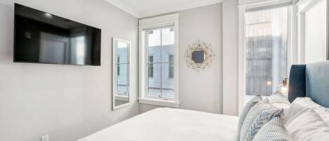 A modern bedroom with a large bed, a flat-screen TV mounted on the wall, and a window with blinds.