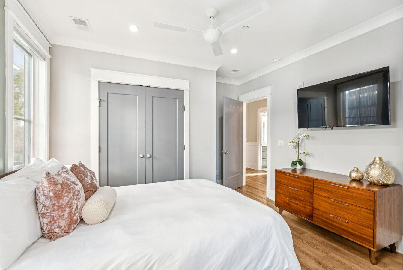 Une chambre avec un grand lit, une armoire grise, une commode en bois et un téléviseur fixé au mur.