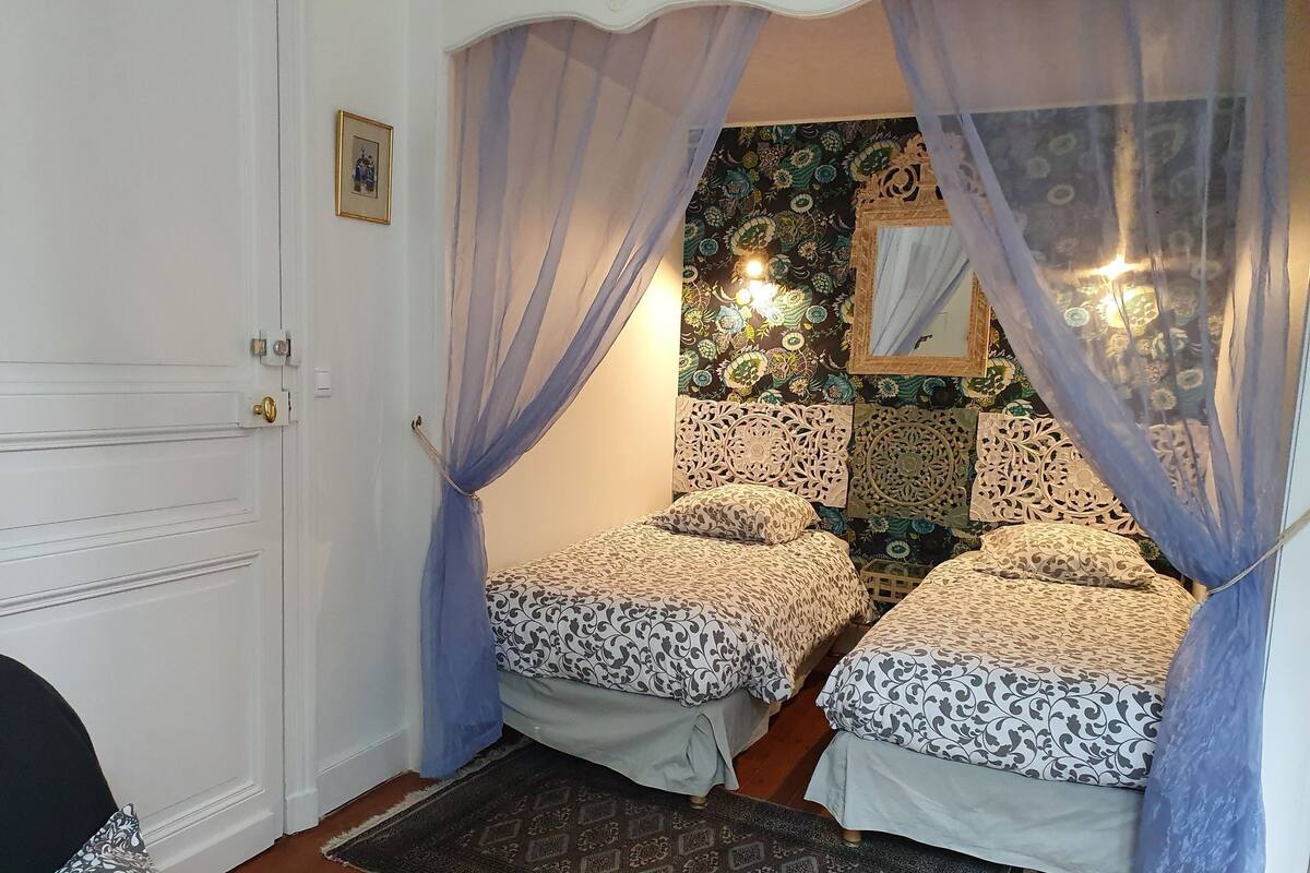 Quarto com duas camas, papel de parede estampado, um candelabro e uma porta com uma pequena imagem.