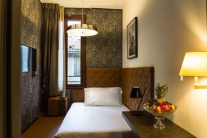 Quarto de hotel com uma cama, uma mesinha de cabeceira com uma fruteira, uma lâmpada e uma televisão.