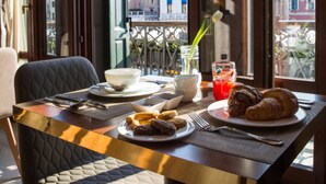 A dining table with breakfast items, a view of buildings through large windows, and a chair.