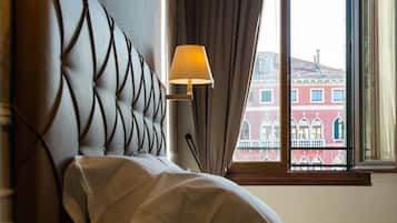 A hotel room with a tufted headboard, a bedside lamp, a clear vase, and a view of buildings through a window.