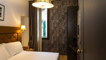A hotel room with a bed, a nightstand, a lamp, a window with curtains, a wall-mounted TV, and a door leading to a bathroom.